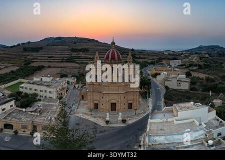 Vue aérienne de l'église paroissiale de Corpus Christi à Gasri, Gozo, avec dôme rouge, façade en calcaire, rues du village et terres agricoles en terrasses au crépuscule. Banque D'Images