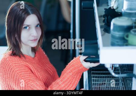 Un jeune barista vêtu d'une tenue orange extrait le porte-filtre de l'équipement espresso dans un café floue. Banque D'Images
