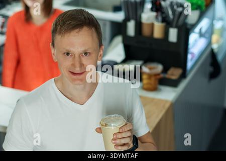Jeune homme souriant en chemise blanche soulève la boisson à emporter dans un café moderne avec un barista en arrière-plan Banque D'Images