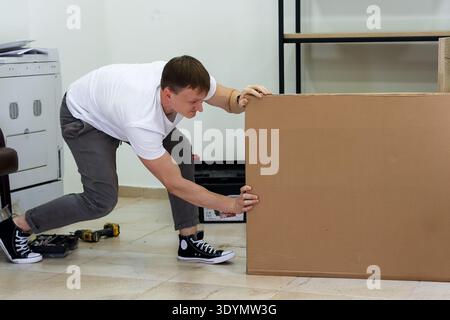 Homme poussant une lourde boîte en carton sur le sol. Homme déterminé en t-shirt blanc poussant une boîte en carton surdimensionnée tout en assemblant des meubles à la maison Banque D'Images