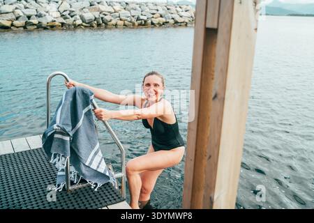 Femme souriante en maillot de bain noir grimpe hors de l'eau sur une terrasse de sauna flottant scandinave. Tenant une serviette, elle respire la joie et la détente, captu Banque D'Images