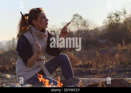 Femme dans la veste décontractée points avec excitation tout en tenant la saucisse sur bâton sur feu de camp incandescent dans la forêt d'automne Banque D'Images