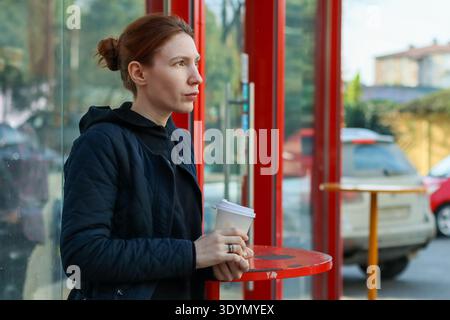 Jeune femme adulte en veste matelassée avec coiffure de pain regarde loin tout en tenant une tasse de café à l'abri d'autobus de la ville pendant la journée Banque D'Images