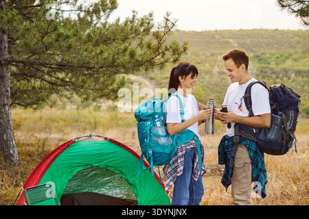 Couple de randonneurs souriants partageant thermos par tente dans la forêt sous le coucher du soleil chaud Banque D'Images