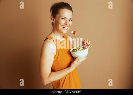 Une femme souriante dans un débardeur jaune et un soutien-gorge de sport regarde la caméra, mangeant une salade saine. Ce studio photographié avec un fond marron clair transporte Banque D'Images