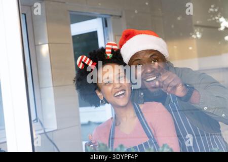 Couple afro-américain debout au comptoir près de la fenêtre, pointant, avec tabliers et coiffures festives Banque D'Images