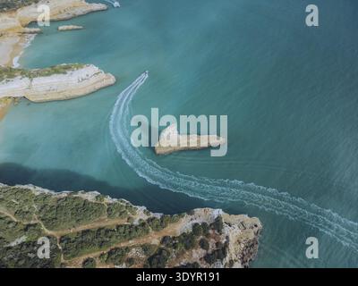 Vue aérienne des eaux turquoises embrassant des falaises accidentées et une formation rocheuse solitaire où les bateaux sculptent des wakes blancs, Porches, Faro, Portugal. Banque D'Images
