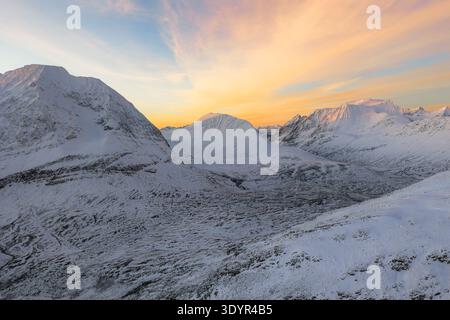 Vue aérienne des montagnes enneigées sous un ciel de coucher de soleil serein, projetant des teintes douces sur le paysage hivernal de Skibotn, Troms, Norvège. Banque D'Images