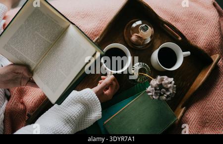 La main tient le livre ouvert, reposant sur une couverture confortable. Deux tasses de café assis sur le plateau en bois à côté. Une petite fleur et un pot en verre complètent la scène. Chaud, poac Banque D'Images