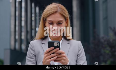 Enthousiaste femme d'affaires caucasienne d'âge moyen heureuse avec le téléphone dans la ville disant wow recevoir la notification réaliser l'opportunité d'emploi femelle étonné Banque D'Images