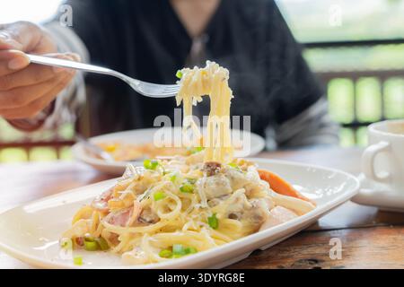 Gros plan de spaghettis servis sur une assiette blanche dans un cadre de repas extérieur avec vue partielle sur une personne tenant une fourchette tout en dégustant un repas Banque D'Images