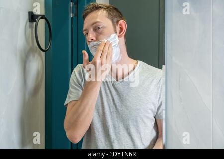 Homme appliquant la mousse de rasage sur le visage dans le miroir de salle de bain. Routine quotidienne de toilettage masculin axée sur le rasage de la barbe et l'hygiène personnelle. Soins personnels et Banque D'Images