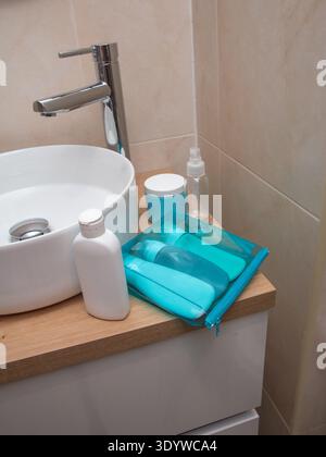 Un ensemble de petites bouteilles rechargeables dans une pochette transparente bleue placée sur un comptoir en bois près d'un évier moderne. Cette image représente Travel préparati Banque D'Images