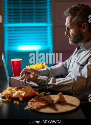Freelance manger de la pizza tard le soir. Bureau de nuit avec ordinateur portable et collation. Détente au bureau de nuit. Travail de nuit. Homme d'affaires mangent collation tout en travaillant sur Banque D'Images