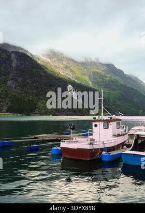 Atmosphère de fjord Tyssedal comme petit bateau de pêche ancré par quai flottant sur l'eau calme du fjord avec des montagnes escarpées et cascade derrière, des coas régionaux Banque D'Images