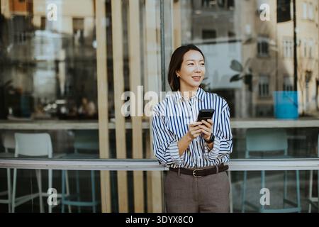 Une coréenne se tient devant un café, regardant son téléphone. Elle porte une chemise rayée et un pantalon gris. L'arrière-plan montre les bâtiments urbains et se Banque D'Images