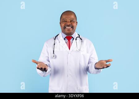Docteur noir mature souriant avec un geste des mains ouvertes sur fond bleu de studio Banque D'Images