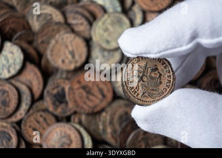 Une photographie en gros plan montre une main dans un gant de protection blanc tenant une ancienne pièce de bronze romaine. La pièce présente un profil détaillé d'un empero Banque D'Images