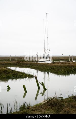 Bateau à voile amarré dans le ruisseau de marée avec des poteaux en bois reflétés dans l'eau à Tollesbury Wick Essex Banque D'Images