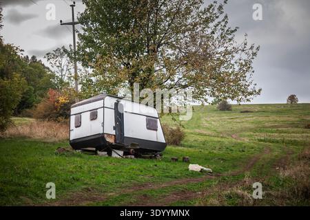 Remorque caravane abandonnée se trouve dans un champ rural herbeux à côté d'un chemin de terre et un arbre solitaire sous un ciel couvert, montrant la négligence et la décomposition dans un co calme Banque D'Images