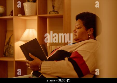 Femme noire d'âge moyen lisant livre tout en étant assise dans le salon confortable Banque D'Images