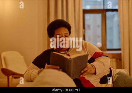 Femme noire d'âge moyen se relaxant à l'intérieur tout en lisant Book on Sofa Banque D'Images