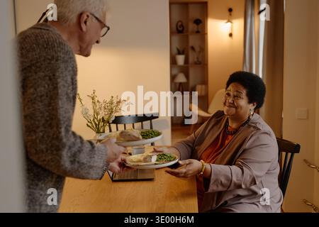 Homme caucasien senior servant le dîner à la femme noire senior à la table à manger Banque D'Images