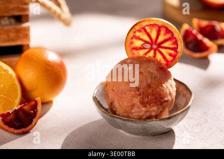 Gros plan d'une boule de sorbet à l'orange sanguine dans un petit bol en céramique, garni d'une tranche d'orange ; entouré de quartiers et d'oranges mûres entières dans un W. Banque D'Images