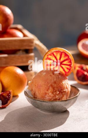 Gros plan d'une boule de sorbet à l'orange sanguine dans un petit bol en céramique, garni d'une tranche d'orange ; entouré de quartiers et d'oranges mûres entières dans un W. Banque D'Images