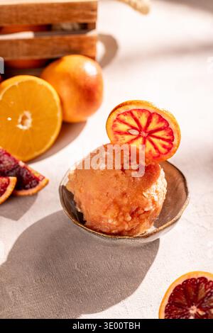 Gros plan d'une boule de sorbet à l'orange sanguine dans un petit bol en céramique, garni d'une tranche d'orange ; entouré de quartiers et d'oranges mûres entières dans un W. Banque D'Images