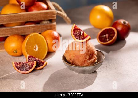 Gros plan d'une boule de sorbet à l'orange sanguine dans un petit bol en céramique, garni d'une tranche d'orange ; entouré de quartiers et d'oranges mûres entières dans un W. Banque D'Images
