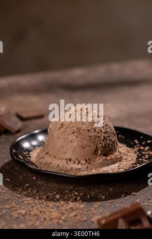 Gros plan d'une grande boule de crème glacée au chocolat crémeuse, saupoudrée de copeaux de chocolat fins, sur une assiette noire mate. Le studio stylé tourné à la guerre Banque D'Images