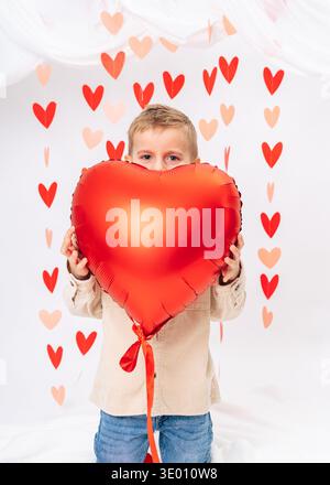 Enfant souriant heureux tenant ballon de coeur rouge sur fond blanc. Saint Valentin, amour, cadeau et concept de célébration d'enfance Banque D'Images