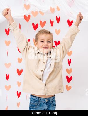 Enfant souriant heureux avec les mains levées entourées de cœurs rouges sur fond blanc. Joie, amour, émotions positives et concept de bonheur infantile Banque D'Images