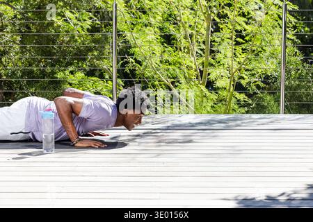 Homme afro-américain effectuant des pompes sur tapis de pont en lavande T, bouteille à proximité, espace copie. Fitness, extérieur, terrasse, lumière du soleil, ombres, verdure, Banque D'Images