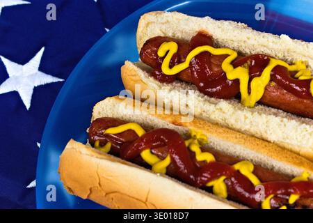 Vue rapprochée de deux hot-dogs américains classiques recouverts de ketchup et de moutarde, présentés sur une plaque bleue sur fond de drapeau américain Banque D'Images