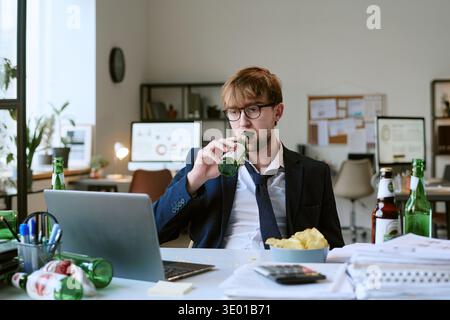 Jeune homme caucasien buvant de la bière tout en travaillant au bureau avec ordinateur portable Banque D'Images
