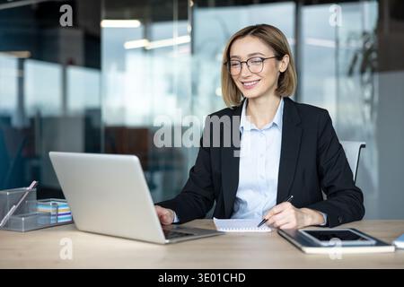 Jeune femme d'affaires dans des lunettes souriant alors qu'elle travaille sur un ordinateur portable, écrit des notes et planifie des tâches dans un bureau moderne, confiant et productif dans un cadre d'entreprise Banque D'Images