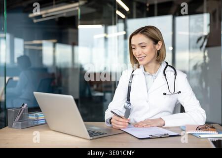 Femme médecin dans un manteau blanc avec un stéthoscope assis à son bureau, écrivant des notes tout en regardant un écran d'ordinateur portable, fournissant une consultation médicale en ligne Banque D'Images