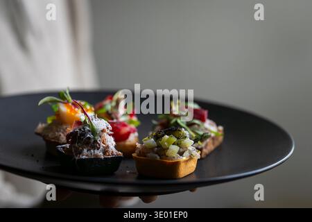 Canapés gastronomiques assortis avec tartare de poisson, fruits de mer et herbes fraîches servis sur une assiette noire dans un cadre gastronomique. Sélection d'entrées élégantes. Banque D'Images