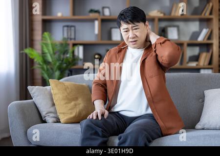 Homme asiatique sur le canapé gris dans le salon frottant mal au cou avec une main, montrant la raideur et l'inconfort d'une mauvaise posture ou d'une fatigue musculaire tout en étant assis à la maison Banque D'Images