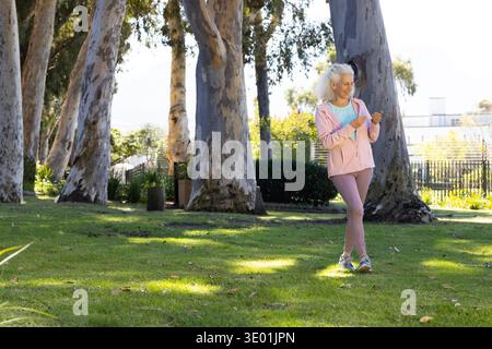 Femme senior en vêtements d'activité debout près du tronc d'eucalyptus à droite du parc tenant le téléphone, espace de copie. Aîné, appareil, extérieur, espace vert, urbain, eucalyp Banque D'Images