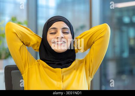 Femme d'affaires musulmane portant un hijab prenant un moment paisible pour se détendre et se déstresser avec les yeux fermés et un sourire satisfait, dans son espace de travail moderne de bureau Banque D'Images