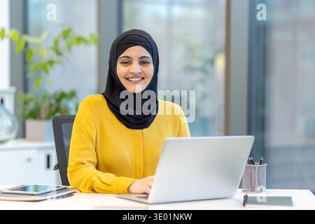 Femme d’affaires musulmane portant un hijab noir et un chemisier jaune, souriant tout en travaillant sur un ordinateur portable à son bureau dans un bureau moderne, représentant le professionnalisme et la diversité sur le lieu de travail Banque D'Images