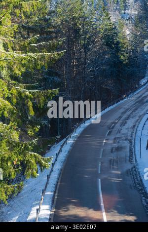 La neige recouvre le sol tandis qu'une route sinueuse serpente à travers une forêt tranquille. La lumière du soir jette une lueur chaude sur le paysage glacé, créant une atmosphère sereine Banque D'Images