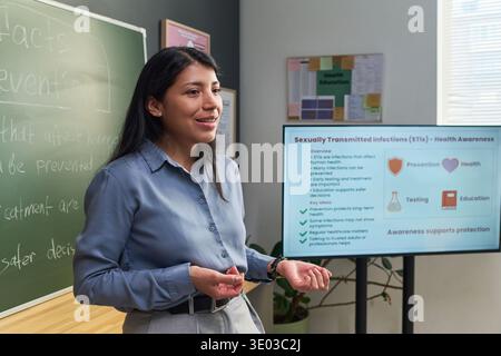 Jeune femme hispanique adulte présentant une leçon d'éducation à la santé sur les infections sexuellement transmissibles, debout dans la salle de classe près du tableau noir et affichage numérique avec des graphiques informatifs Banque D'Images