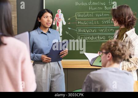 Jeune femme hispanique adulte expliquant les systèmes du corps humain à un groupe d'adolescents en classe, tenant une presse-papiers, debout devant un tableau avec un diagramme anatomique, les étudiants écoutant attentivement Banque D'Images