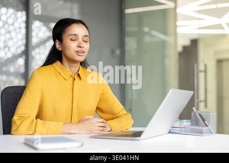 Jeune femme fermant les yeux, pratiquant la pleine conscience et la respiration profonde à son bureau, trouvant la paix intérieure tout en prenant une pause du travail pour gérer le stress et l'anxiété Banque D'Images