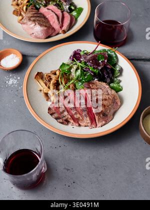 Le steak tendre de chuck tranché est présenté avec une salade verte fraîche et des champignons. Deux verres de vin rouge sont assis à proximité sur une surface de pierre sombre, créant un Banque D'Images