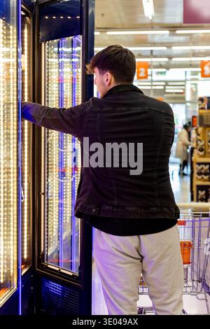 Un jeune homme portant une veste noire et un pantalon gris sélectionne des produits dans une section réfrigérée d'une épicerie, avec un chariot à proximité Banque D'Images